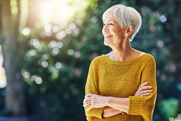 A woman in a yellow sweater crosses her arms and stares in the distance longingly. She's outside in a garden.