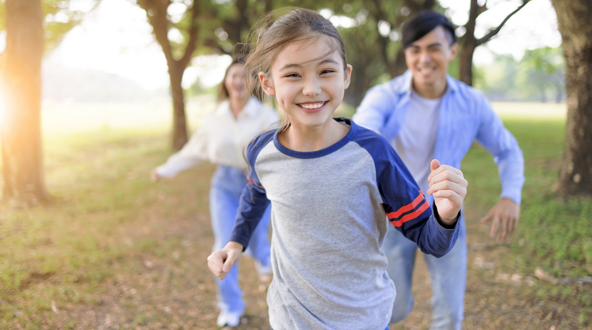 Young girl running outside in the park with her parents, enjoying playtime and exercise.