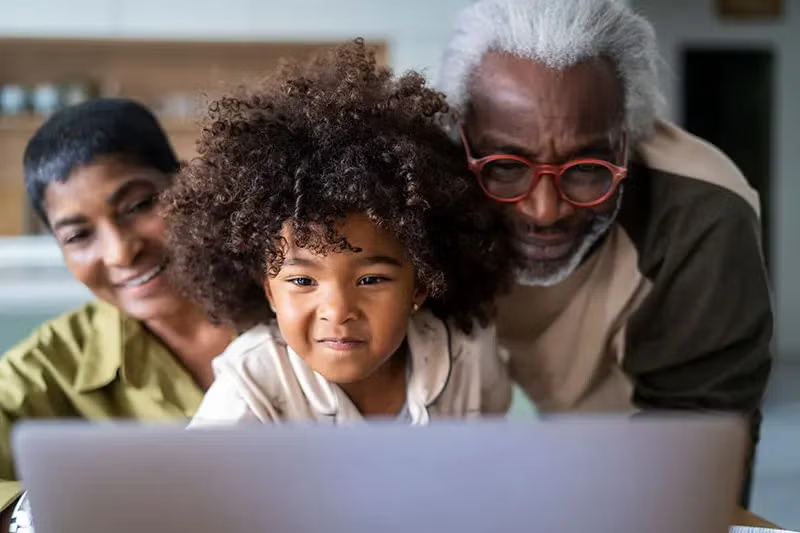 Two grandparents stand behind their young grandaughter as they all look at a laptop screen